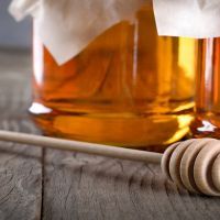 Pouring aromatic honey into jar, closeup. Honey in glass jars and honeycombs wax on wooden background. Wooden stick , instruments